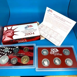 May include: A 2004 United States Mint Silver Proof Set. The set includes a box with the Statue of Liberty, a certificate, and two clear cases containing silver coins. The cases display various coins, including quarters and half dollars.
