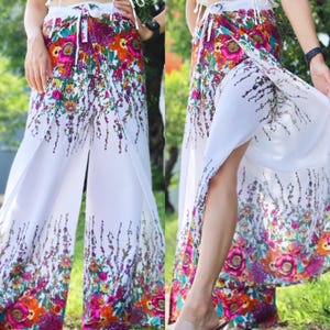 May include: White palazzo pants with a vibrant floral print in shades of pink, orange, and purple. The pants feature a split-leg design and a drawstring waist. The floral pattern is concentrated at the top and bottom of the pants.