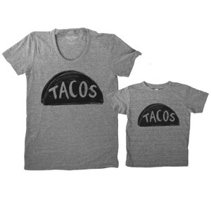 May include: Two grey t-shirts with a black graphic of a taco shell with the word "TACOS" written inside. The larger shirt is for adults and the smaller shirt is for children.