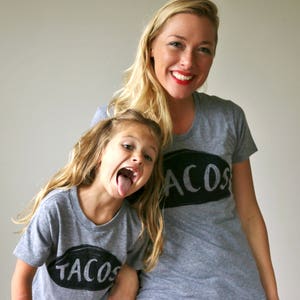 May include: A mother and daughter wearing matching grey t-shirts with the words "TACOS" and "ACOS" printed on them. The daughter is sticking her tongue out.