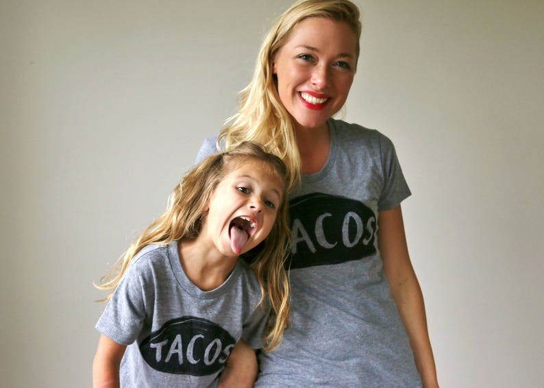 May include: A mother and daughter wearing matching grey t-shirts with the words "TACOS" and "ACOS" printed on them. The daughter is sticking her tongue out.