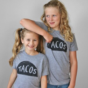 May include: Two girls wearing grey t-shirts with the words "Tacos" and "Acos" printed on them. The girls are smiling and looking at the camera.