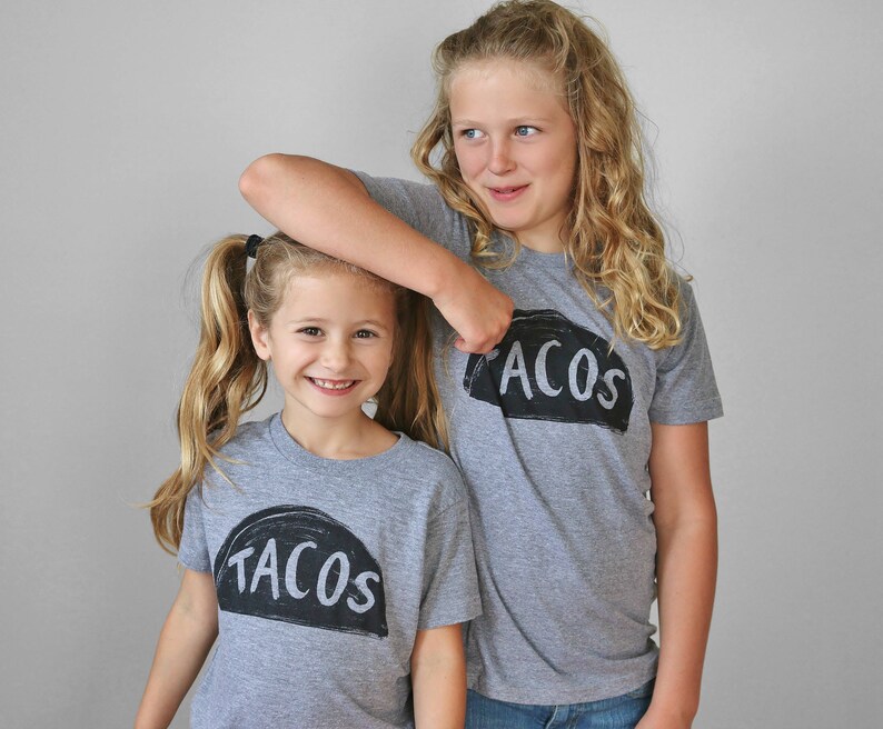 May include: Two girls wearing grey t-shirts with the words "Tacos" and "Acos" printed on them. The girls are smiling and looking at the camera.