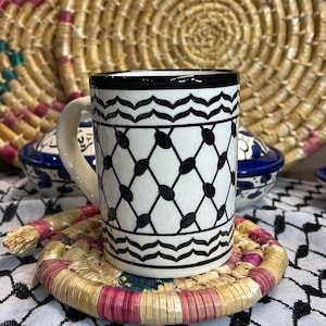 May include: A white ceramic mug with a black geometric pattern and a matching coaster. The mug has a curved handle and a black rim. The coaster is made of woven straw with pink and gold accents. The background includes woven baskets.