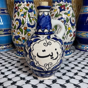 May include: A blue and white ceramic oil bottle with intricate floral designs and Arabic calligraphy. The bottle has a cork stopper and a handle. The bottle is sitting on a white and black checkered surface.