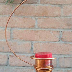 May include: A copper wire hummingbird feeder with a red plastic top hanging from a branch.