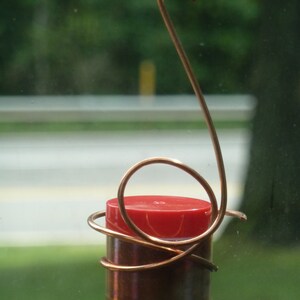 May include: A red and copper colored glass bottle with a copper wire hanger. The bottle is hanging from a window.