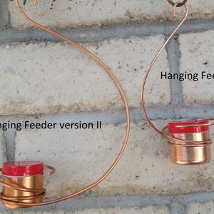 Pode incluir: Dois comedouros para p&aacute;ssaros suspensos de arame de cobre com tampas vermelhas. O comedouro &agrave; esquerda est&aacute; etiquetado como "Hanging Feeder version II" e o comedouro &agrave; direita est&aacute; etiquetado como "Hanging Feeder version I".