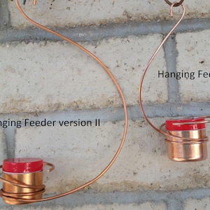 May include: Two copper wire hanging bird feeders with red lids. The feeder on the left is labeled "Hanging Feeder version II" and the feeder on the right is labeled "Hanging Feeder version I".