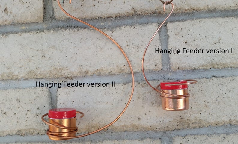 May include: Two copper wire hanging bird feeders with red lids. The feeder on the left is labeled "Hanging Feeder version II" and the feeder on the right is labeled "Hanging Feeder version I".