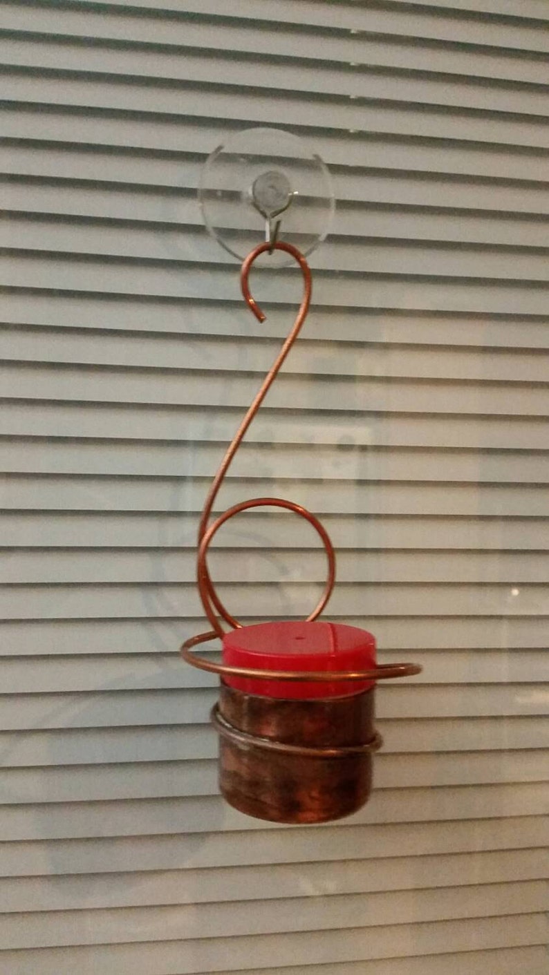 May include: A copper hummingbird feeder with a red top, hanging from a suction cup on a window.