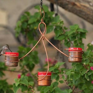May include: A copper hummingbird feeder with three red feeding stations. A hummingbird is drinking nectar from one of the feeders.