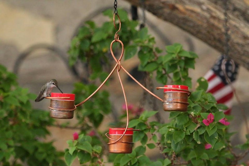 May include: A copper hummingbird feeder with three red feeding stations. A hummingbird is drinking nectar from one of the feeders.