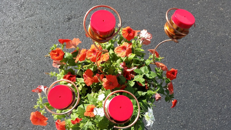 May include: A copper hummingbird feeder with four red lids and spiral accents, surrounded by vibrant orange and red flowers and green foliage. The feeder is designed to attract hummingbirds and adds a decorative touch to any garden.