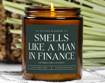 May include: A brown glass candle jar with a green label that reads "LETTUCE BLOSSOM SMELLS LIKE A MAN IN FINANCE CONGRATULATIONS! DYLAN HAWK, MBA ESTABLISHED 2024".
