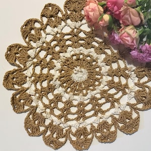 May include: A handmade, circular crochet doily in shades of brown and white. The doily features a detailed, openwork design with a scalloped edge. A bouquet of pink and purple roses rests beside the doily.