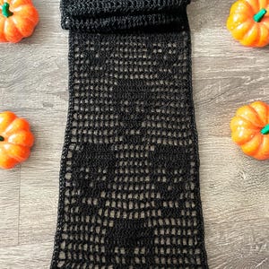 May include: A long, rectangular black crochet runner featuring an openwork mesh pattern that forms a repeating skull motif. The fabric is constructed with thin black yarn, creating a semi-sheer, grid-like texture throughout the entire length.