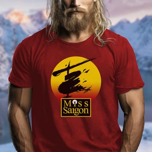 以下が含まれることがあります： 赤色のTシャツ。黄色とオレンジ色の円形デザインで、ヘリコプターと顔の黒いシルエットが描かれています。「Miss Saigon」の文字は、黒い縁取りのある黄色のボックス内にあります。