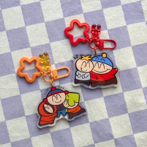 Peut inclure: Trois porte-clés avec des personnages de dessins animés. L'un a une étoile orange, un autre une étoile rouge, et le troisième représente trois personnages qui s'embrassent. Chaque porte-clés comprend un fermoir.