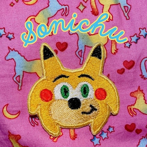 May include: A yellow embroidered patch of a cartoon character with green eyes and a smirk, on a pink fabric background with a rainbow unicorn pattern and the text "sonicchu" in yellow and blue.