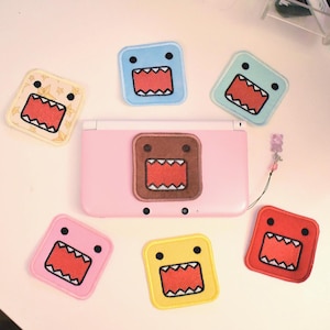 May include: A pink Nintendo DS console with a brown Domo-kun patch, surrounded by six colorful Domo-kun patches. The patches are square with a red mouth and black eyes, in various colors including blue, pink, and yellow.