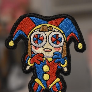 May include: Embroidered patch featuring a cartoon jester character. The jester has a red, blue, and yellow color scheme, with a black outline. The jester's eyes have a unique design, and the patch is likely for clothing or accessories.