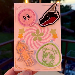 May include: Four embroidered iron-on patches featuring a pink character with a surprised expression, a red and black dragon, a yellow character with a star, and a green character with a surprised expression. The patches are on a white card with a pink and green swirl design.