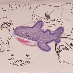 May include: A purple shark patch with a pink mouth and white teeth is centered on a white background with the word "BLÅHAJ" above it. The image also includes several sketched shark illustrations in black ink.