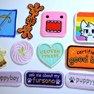 May include: A collection of embroidered patches in various shapes and designs. Includes a scale of justice, a Domo-kun character, a cat with a rainbow background, and patches with text like "certified good boy" and "puppyboy".