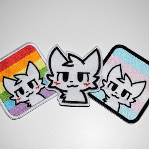 May include: Three embroidered patches featuring a white cat-like character with black eyes and a red blush. One patch has a rainbow background, another has a transgender flag background, and the third is a standalone character.