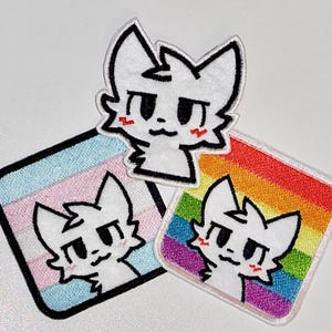 May include: Three embroidered patches featuring a white cat-like character with black outlines and red cheeks. One patch is a cat head, another is square with a transgender flag, and the third is square with a rainbow.