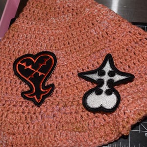 May include: A peach-colored crocheted hat with two embroidered patches. One patch is a black heart with a red outline and a broken design. The other is a white and black star-shaped patch. The hat is made of a soft, textured yarn.