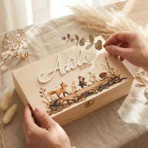 Peut inclure: Une boîte en bois de couleur claire avec un couvercle à charnière. La boîte est décorée d'une scène de forêt avec un renard, un lapin, un raton laveur et un hérisson. Le nom "Adele" est écrit sur le dessus, avec la date "23.05.2026".