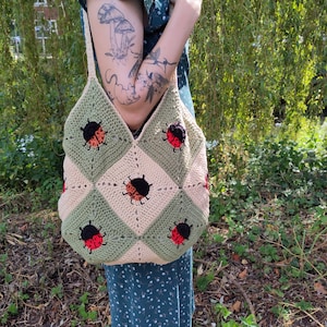 Op de afbeelding: Een gehaakte tote bag met een groen en beige ruitpatroon en lieveheersbeestje-applicaties. De tas wordt gedragen door een persoon met een tatoeage op hun arm.