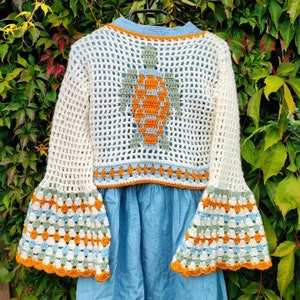 Op de afbeelding: Een wit gehaakt topje met een schildpadontwerp op de rug. Het topje heeft wijde mouwen met een meerkleurige gehaakte rand. Het topje wordt gedragen over een lichtblauwe denimjurk.