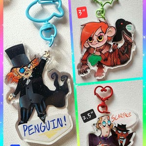 Könnte beinhalten: Vier Schlüsselanhänger aus Acryl mit Cartoon-Charakter-Designs. Ein Schlüsselanhänger zeigt einen Charakter mit dem Wort "PENGUIN!" und ist 7,6 cm groß. Ein weiterer Schlüsselanhänger hat einen Charakter mit dem Text "SCARFACE and ARNOLD WESKER" und ist 6,4 cm groß. Die anderen zwei haben einzigartige Designs.