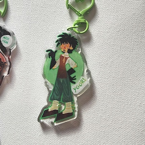 May include: A clear acrylic keychain featuring a cartoon character with dark hair, glasses, and a brown vest. The character is set against a green background with the word "YGOR!" and attached to a green heart-shaped clasp.