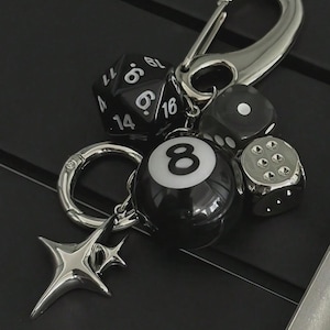 May include: A silver-toned keychain featuring a variety of charms. Included are black dice, a black and white 8-ball, and a star-shaped charm. The keychain has a large clasp for easy attachment.