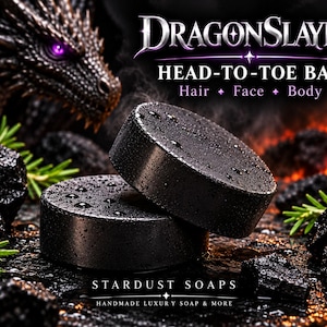 May include: Two round black soap bars with water droplets on the surface. The text reads DRAGONSLAYER HEAD-TO-TOE BAR Hair Face Body and STARDUST SOAPS HANDMADE LUXURY SOAP & MORE. The background features a dragon head, charcoal, and green sprigs.