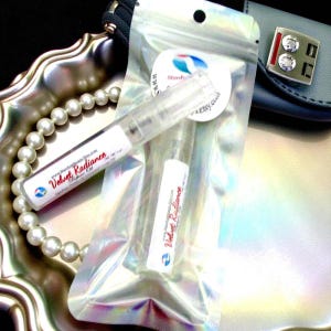 May include: Clear perfume vials in a holographic zip bag. One vial is labeled "Velvet Radiance" with a website address. A pearl necklace and a silver tray are in the background. A sticker with a logo and website address is also visible.