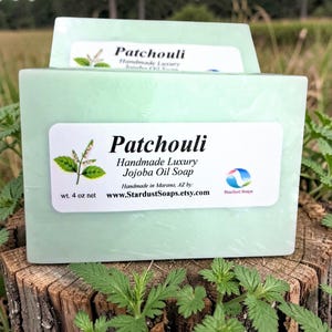 Peut inclure: Un pain de savon Patchouli vert clair avec une étiquette blanche. L'étiquette indique "Patchouli Handmade Luxury Jojoba Oil Soap" et "wt. 4 oz net". Le savon est présenté sur une surface en bois.