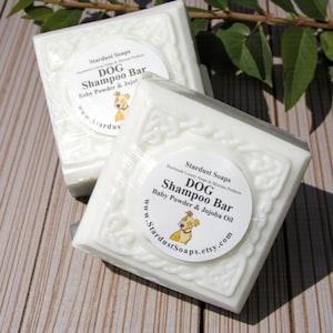 May include: Two white dog shampoo bars with decorative edges are displayed. Each bar has a circular label with the text "DOG Shampoo Bar" and "Baby Powder & Jojoba Oil". The bars are from Stardust Soaps.