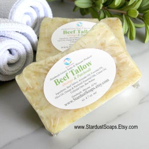 Puede incluir: Tres barras de jabón hechas a mano, envueltas en plástico transparente, con etiquetas ovaladas. Las etiquetas dicen "Beef Tallow Orange & Patchouli" y enumeran los ingredientes. El jabón es de color amarillo claro con motas visibles.