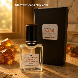 May include: A clear glass 15ml perfume bottle with a black cap and a white label reading Stardust Soaps AMBER EAU DE PARFUM 15ml StardustSoaps.etsy.com, positioned next to a matching black rectangular box with the same label text.