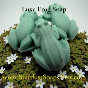 May include: Three light green frog-shaped soaps are arranged on a bed of green foliage with small white flowers. The text "Luxe Frog Soap" is at the top, and "www.StardustSoaps.Etsy.com" is at the bottom.