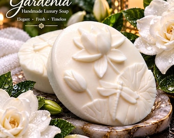 Może przedstawiać: Dwie okrągłe, kremowobiałe kostki mydła z wytłoczonymi wzorami kwiatu lotosu, ważki i pąka. Widoczny jest tekst "Gardenia Handmade Luxury Soap Elegant Fresh Timeless" oraz "Stardust Soaps". Mydło spoczywa na marmurowej podstawce.