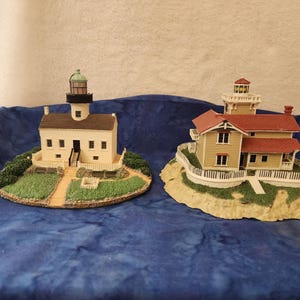 May include: Two miniature lighthouse figurines. One is white with a black tower and green top, the other is yellow with a red roof. Both are set on small, grassy bases with detailed landscaping.