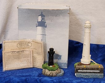 May include: A set of two lighthouse figurines, one white and one black, with a box and a card that says "Harbour Lights." The white lighthouse is on a base with a brown wooden structure and a blue water base. The black lighthouse is on a green base with a brown wooden structure. The card has information about the "Panama Canal Marshal Set."