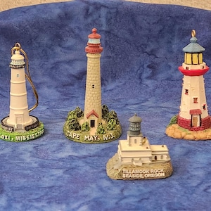 May include: Four miniature lighthouse figurines. One is white with a gold loop, another is beige with a red top, and a third is white with a red and blue top. The fourth is a small white building with a gray top.