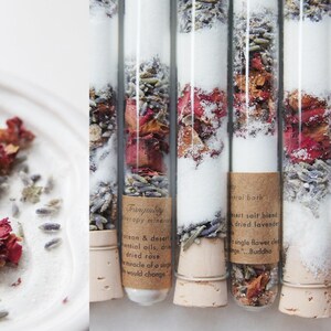 May include: Clear glass tubes filled with white granules, dried lavender, and rose petals. Each tube has a cork stopper and a brown label with text. The tubes are arranged vertically, suggesting a spa or bath product.